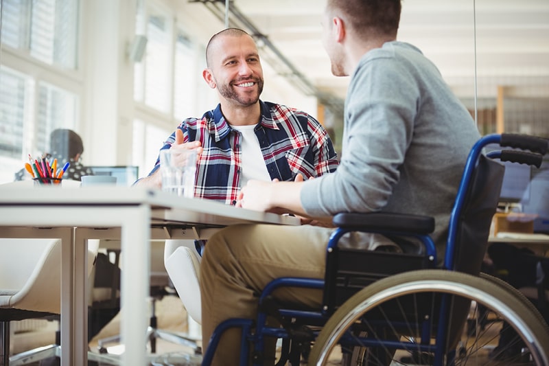 A man in a wheelchair sits at a desk talking to a colleague