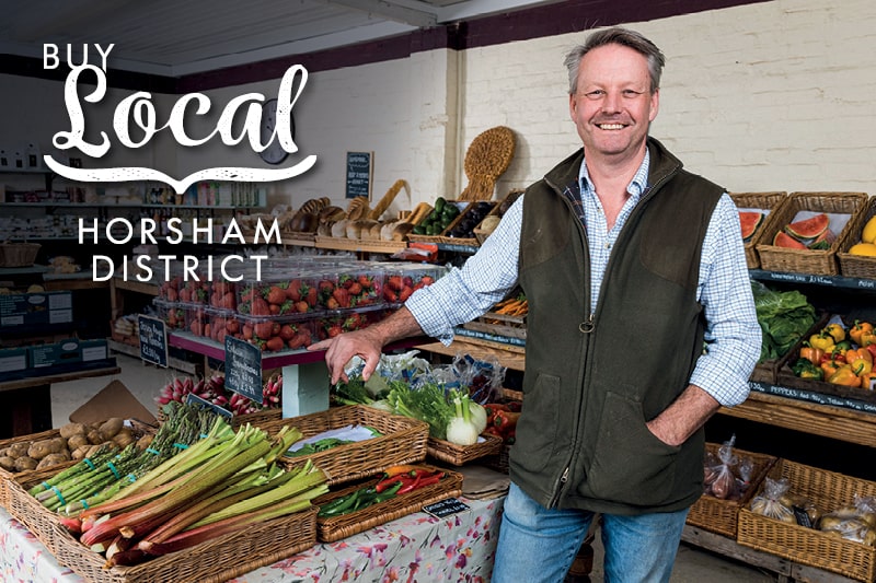 A local shop owner stands smiling with fruit and vegetables. The Buy Local Horsham District logo is in the top corner of the image